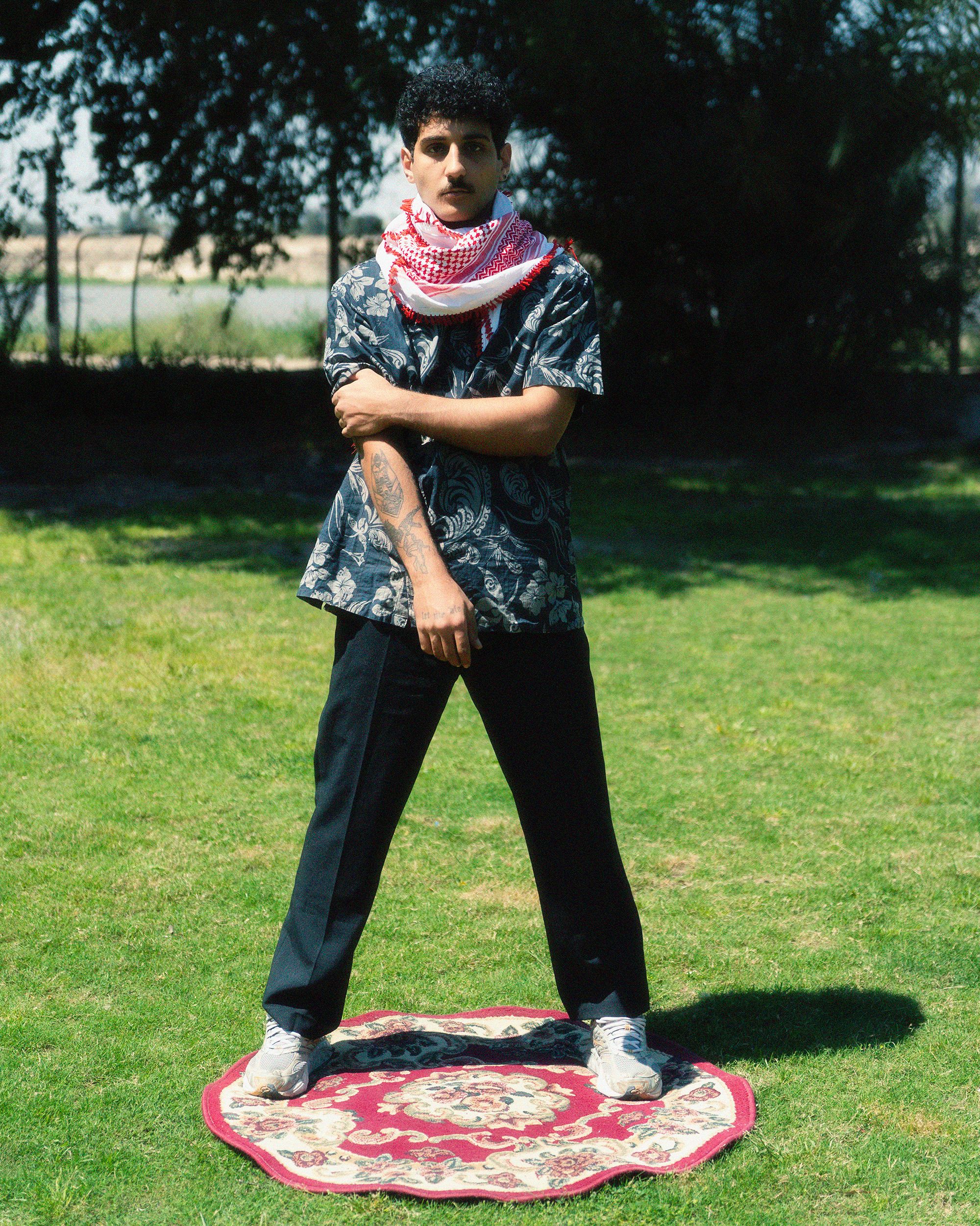 A man in fashionable attire stands confidently outdoors on a patterned rug.