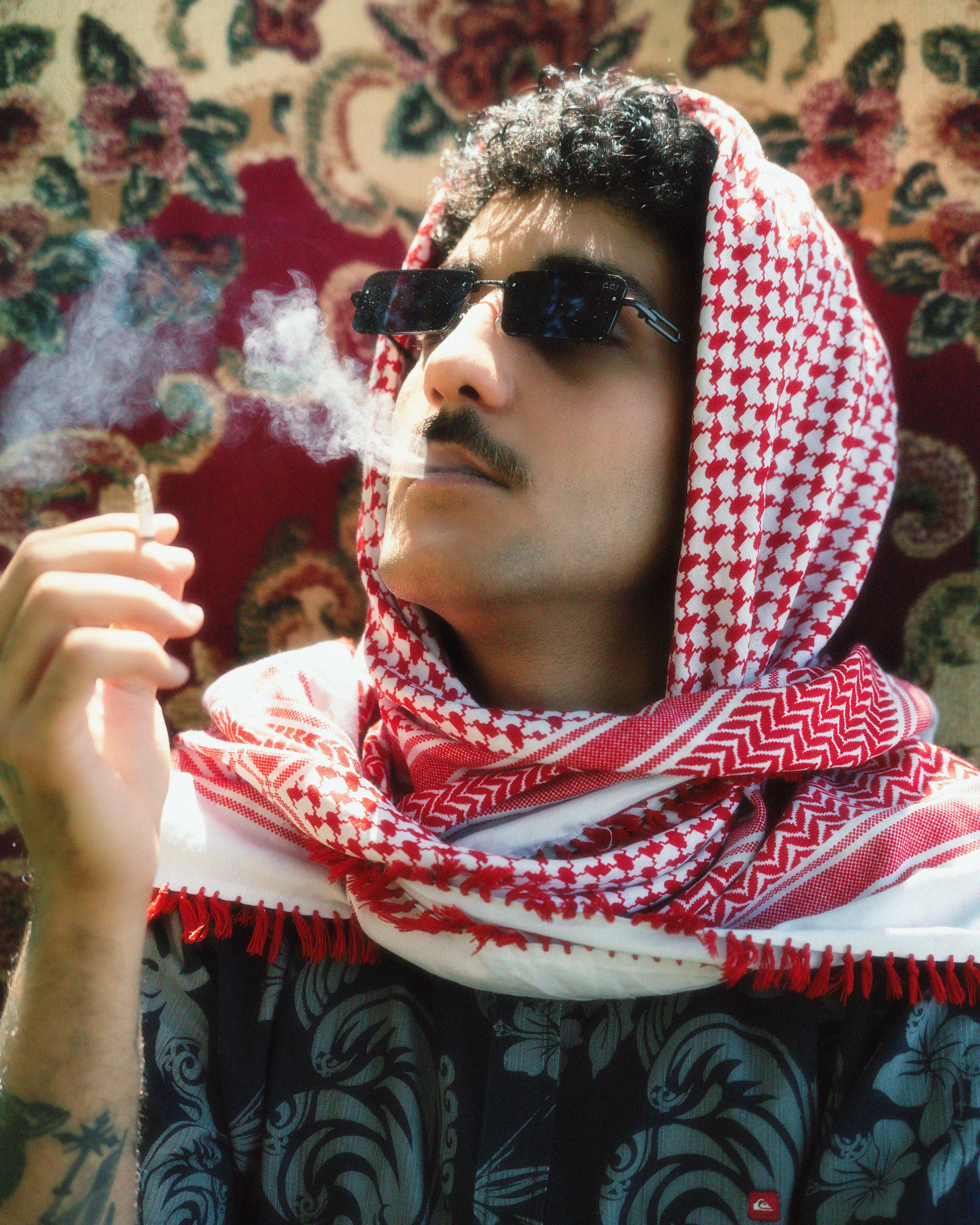Portrait of a man in sunglasses smoking while wearing a red and white shawl, exuding fashion and attitude.