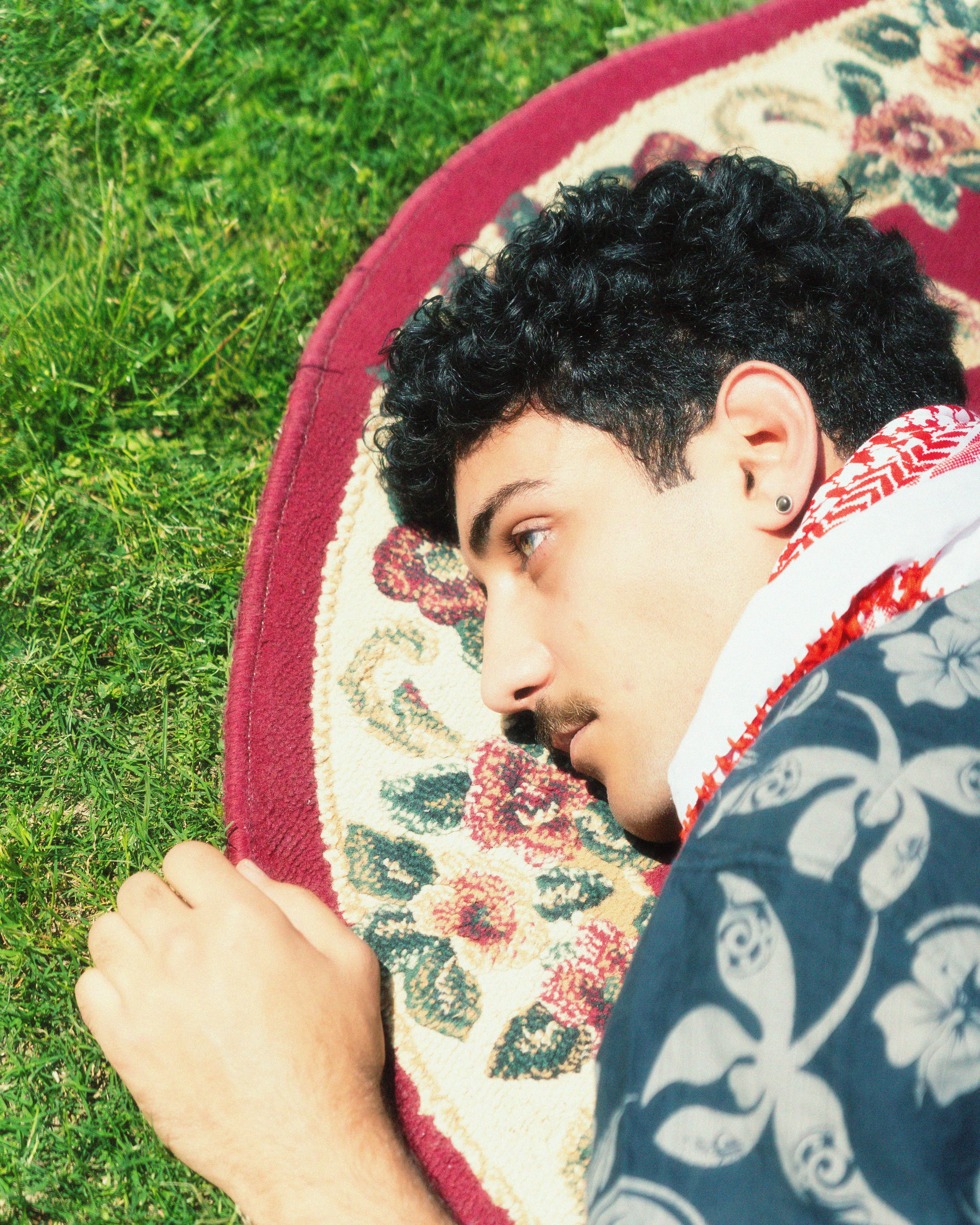 A man with curly hair lying on a floral carpet outside, enjoying a sunny day.