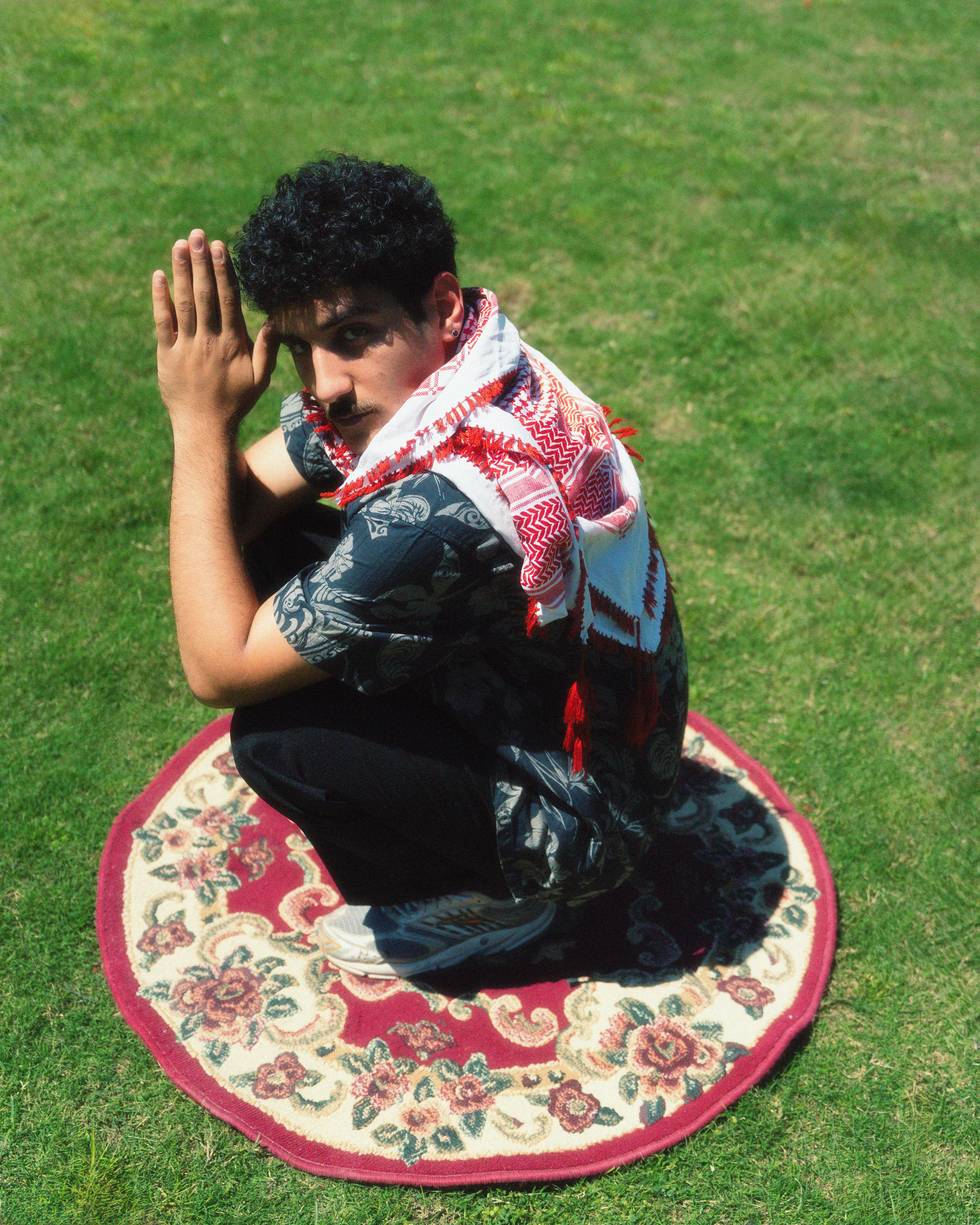 Man squatting on a rug outdoors with hands folded, in a thoughtful pose.