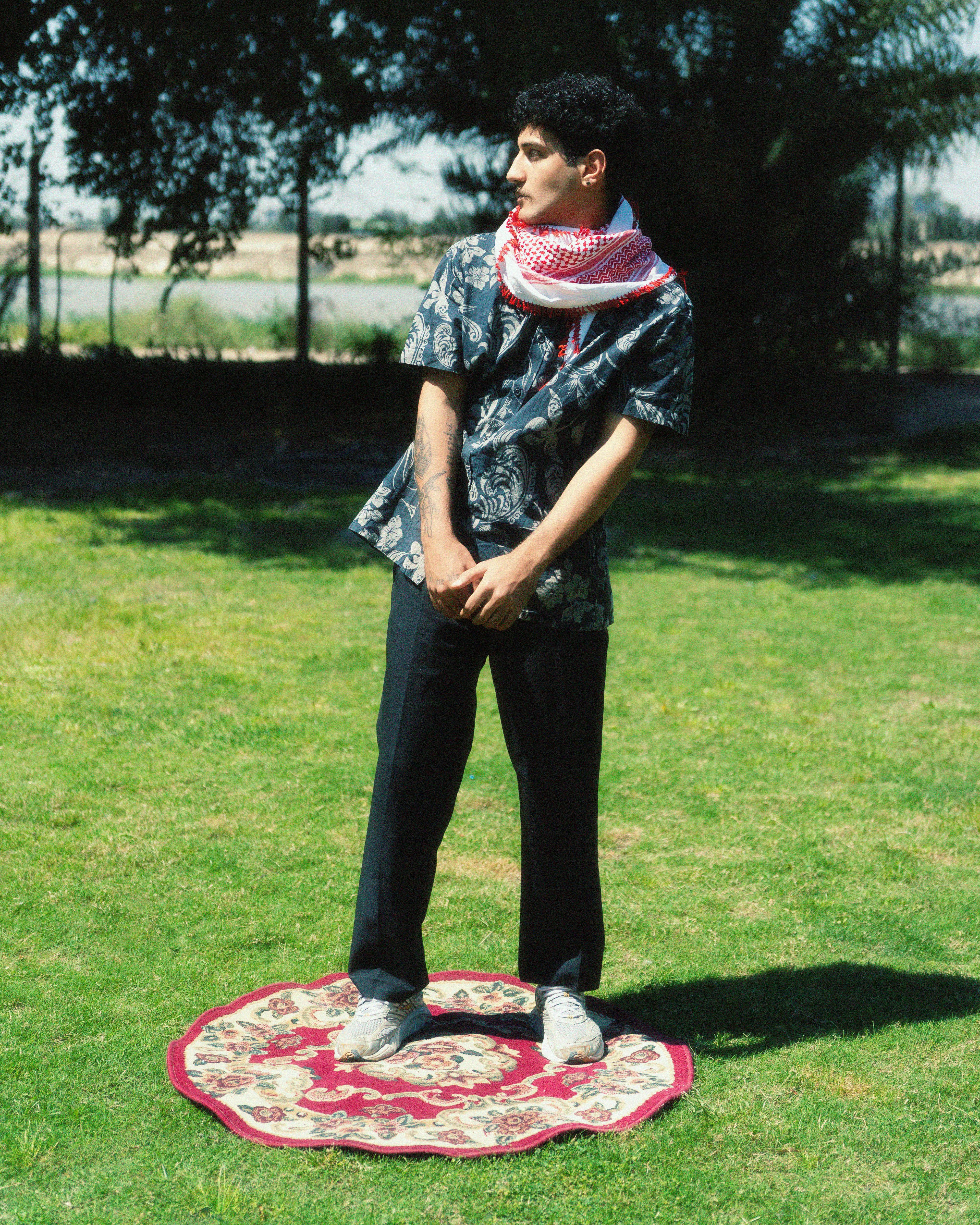 A man in patterned attire stands on a rug in a sunny park, surrounded by green grass and trees.