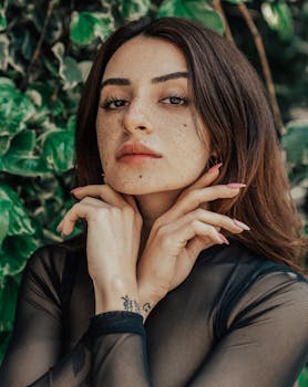 Portrait of a young woman with freckles and tattoos posing outdoors against a green leafy background.