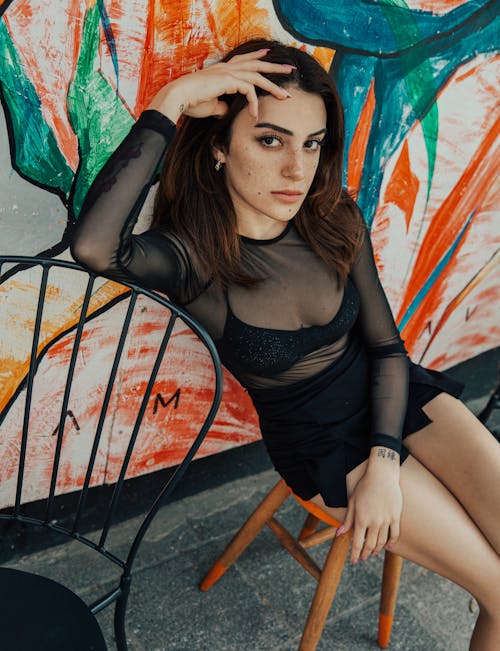 Young Woman in a Black Mesh Blouse and Miniskirt Sitting by a Colorful Wall 