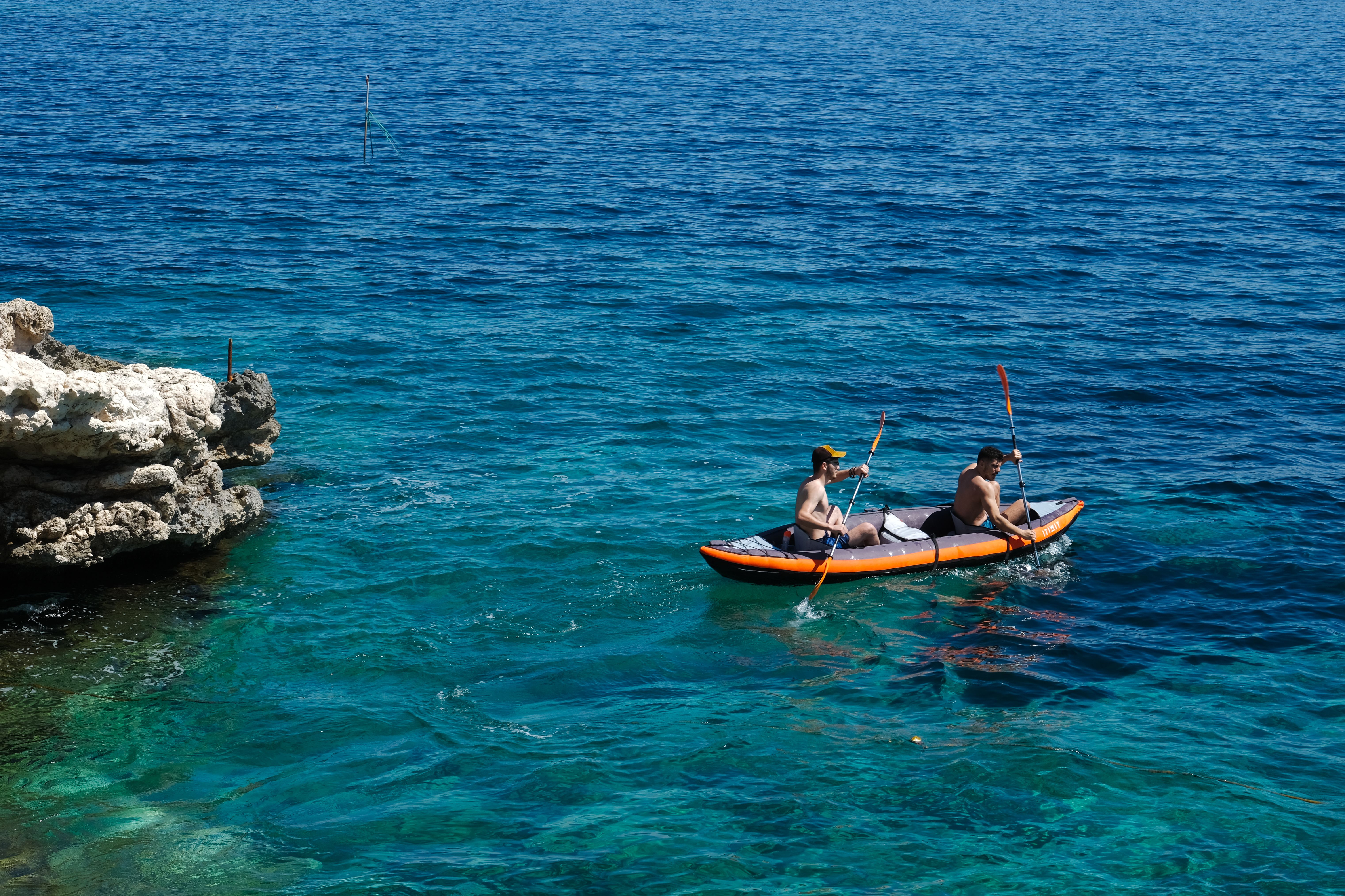 Two men enjoy kayaking in the crystal-clear sea near a rocky shore under the summer sun.