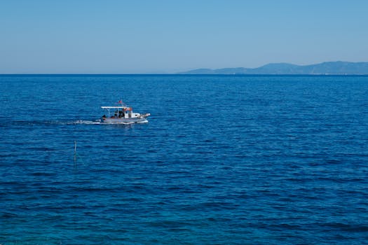 A small boat sails peacefully across a vast blue ocean under a clear sky, with distant hills in view.