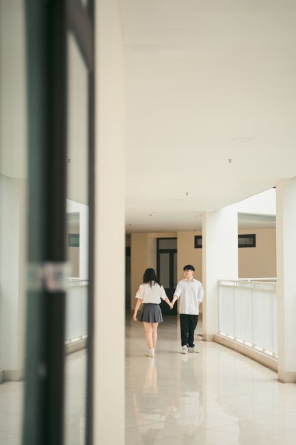 Asian couple walking hand in hand through a bright corridor, showcasing contemporary fashion.