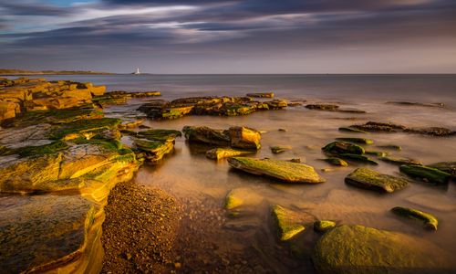 Free A tranquil coastal scene at Whitley Bay with rocky formations and a distant lighthouse during sunset. Stock Photo