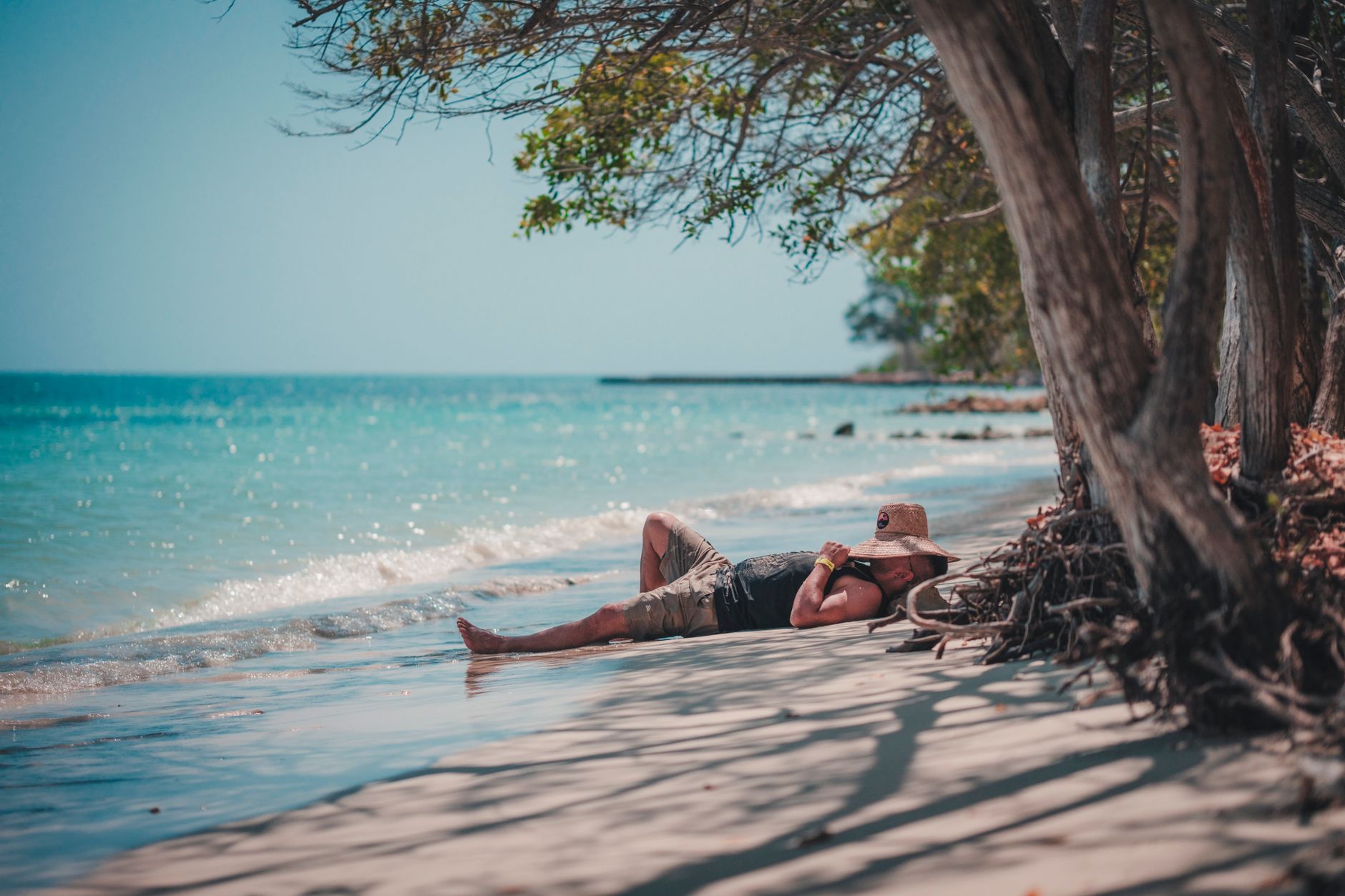 A man in a hat enjoys a tranquil summer day on a tropical beach under the shade of trees.