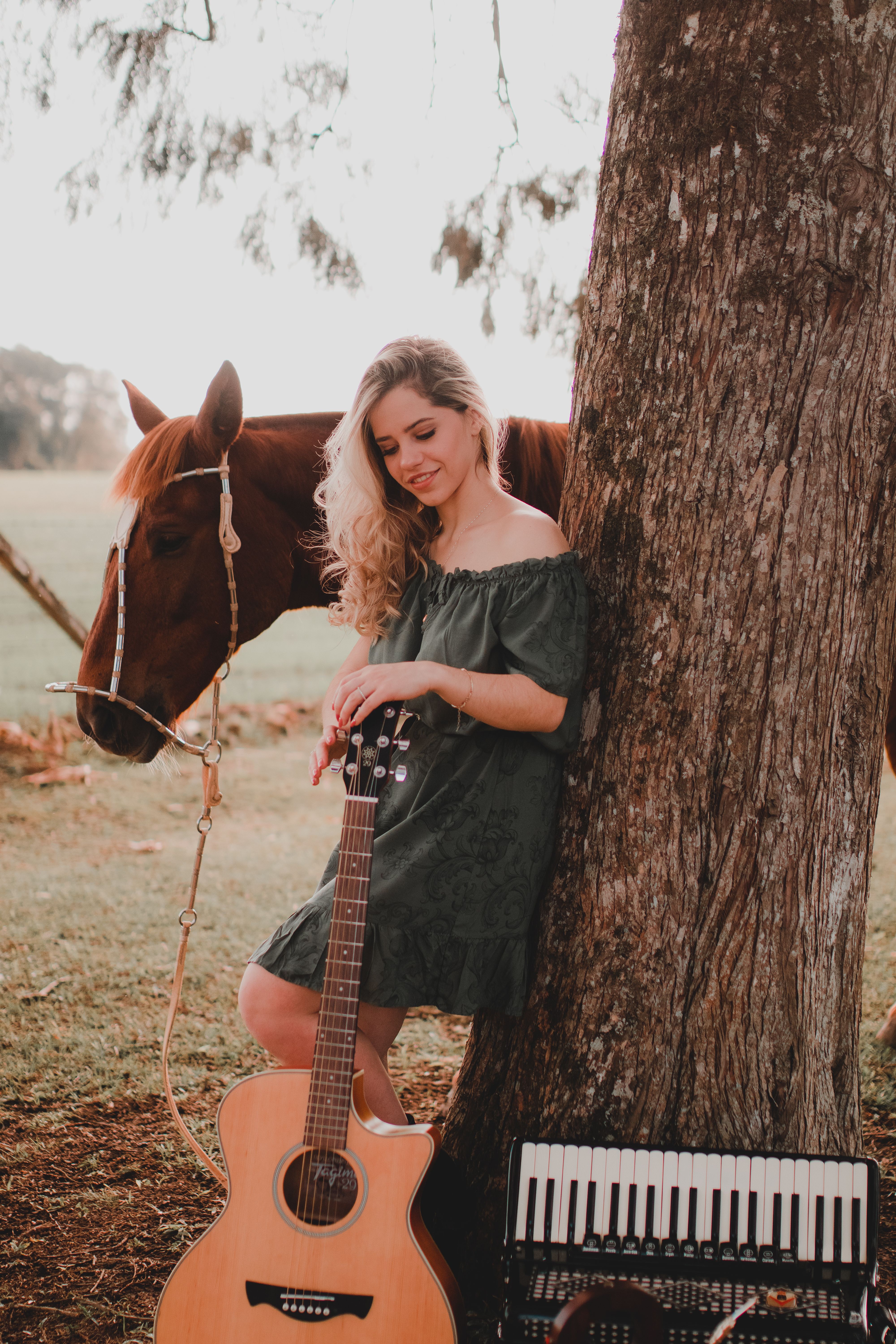 Woman in sundress with guitar and horse outdoors in a rural setting.