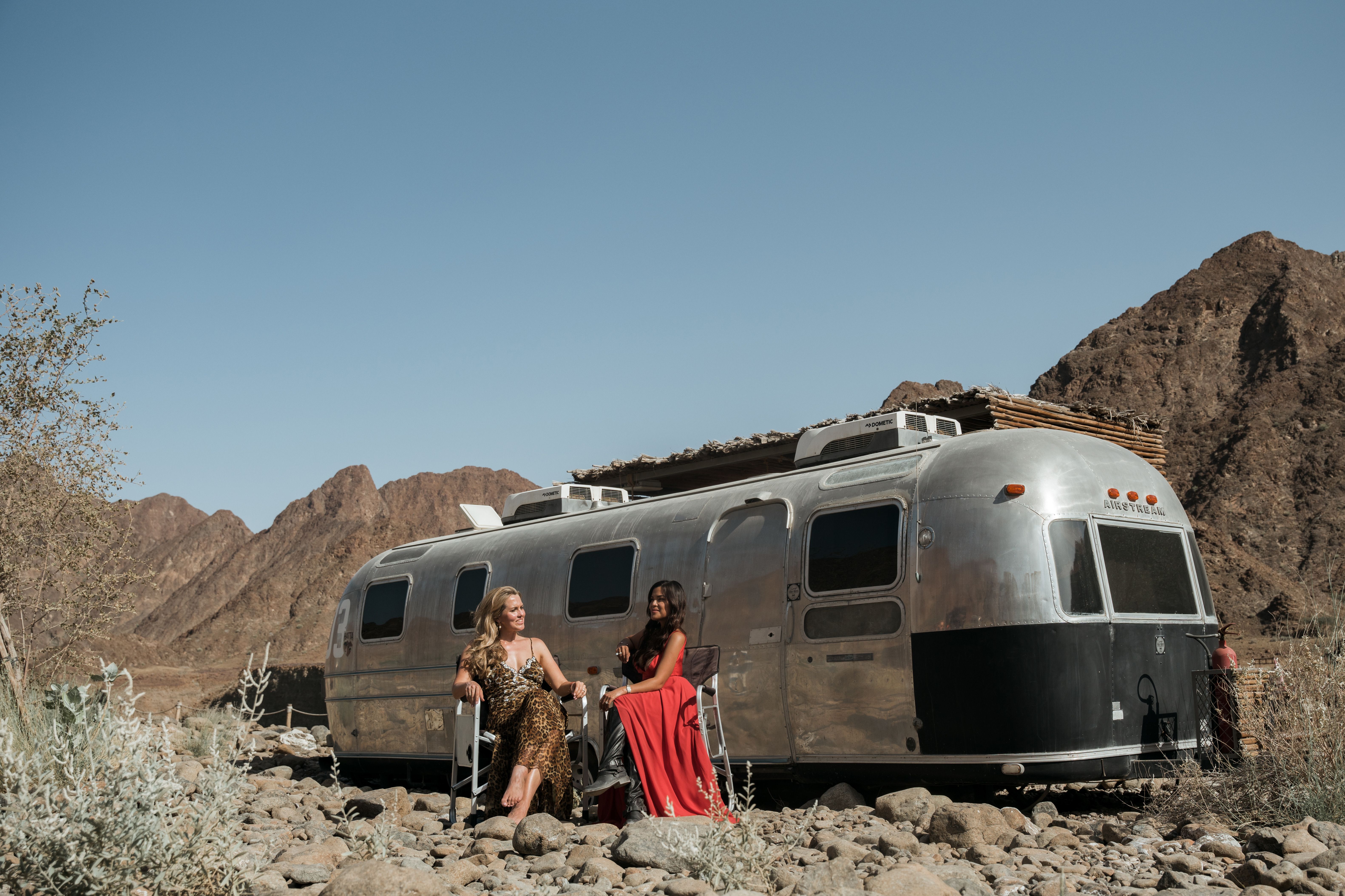 Two women enjoying the summer in Hatta, Dubai with a classic camper and mountainous desert backdrop.