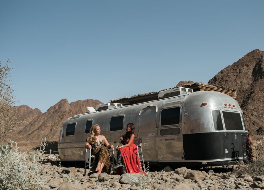 Two women enjoying the summer in Hatta, Dubai with a classic camper and mountainous desert backdrop.