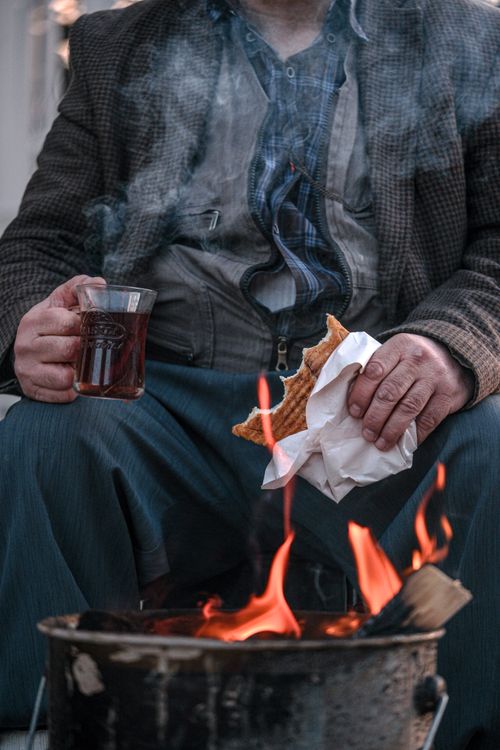 Free A man sits by a warm campfire holding tea and a sandwich, perfect for relaxation. Stock Photo