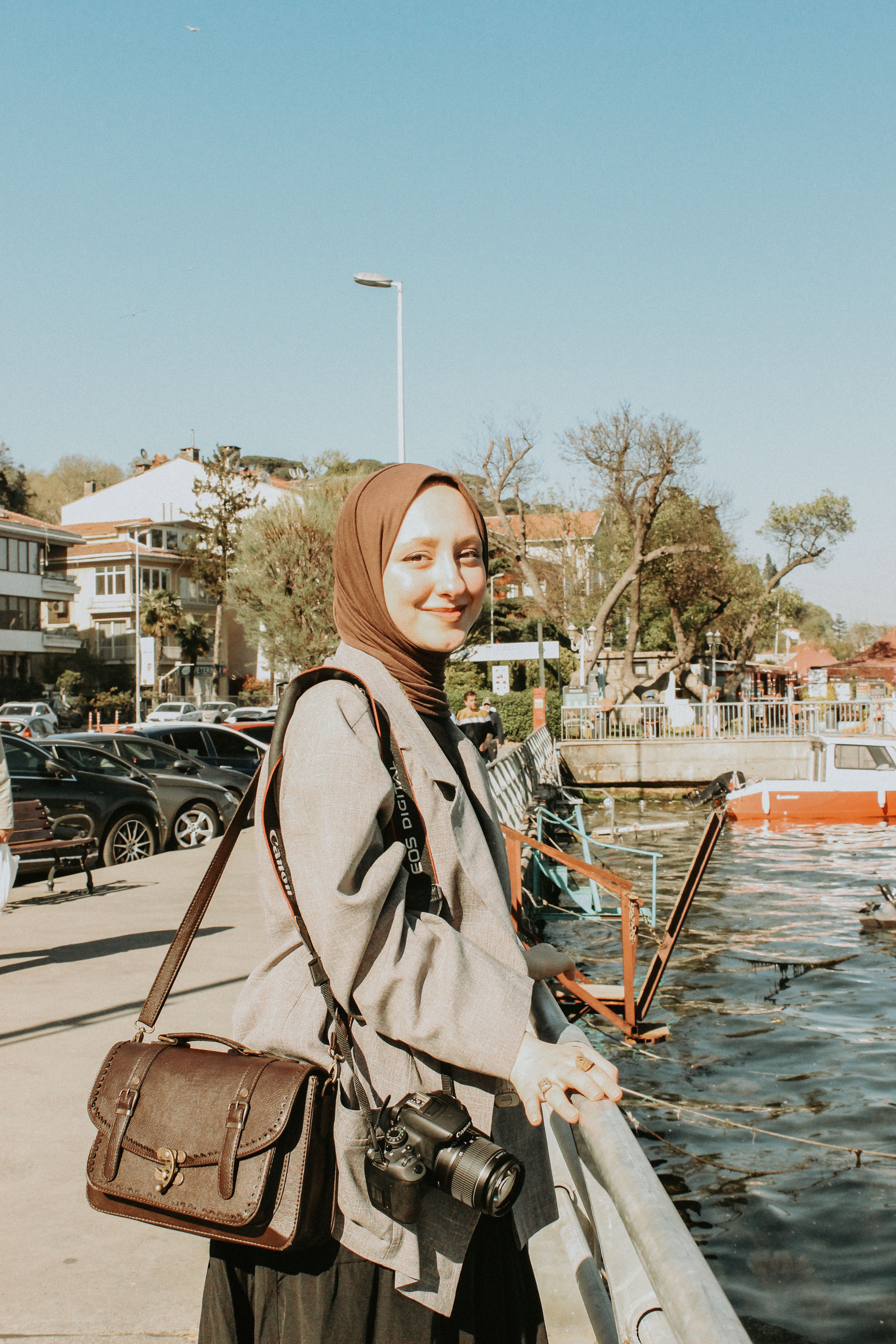A smiling young woman wearing a hijab stands by a city waterfront, enjoying the sunny day.