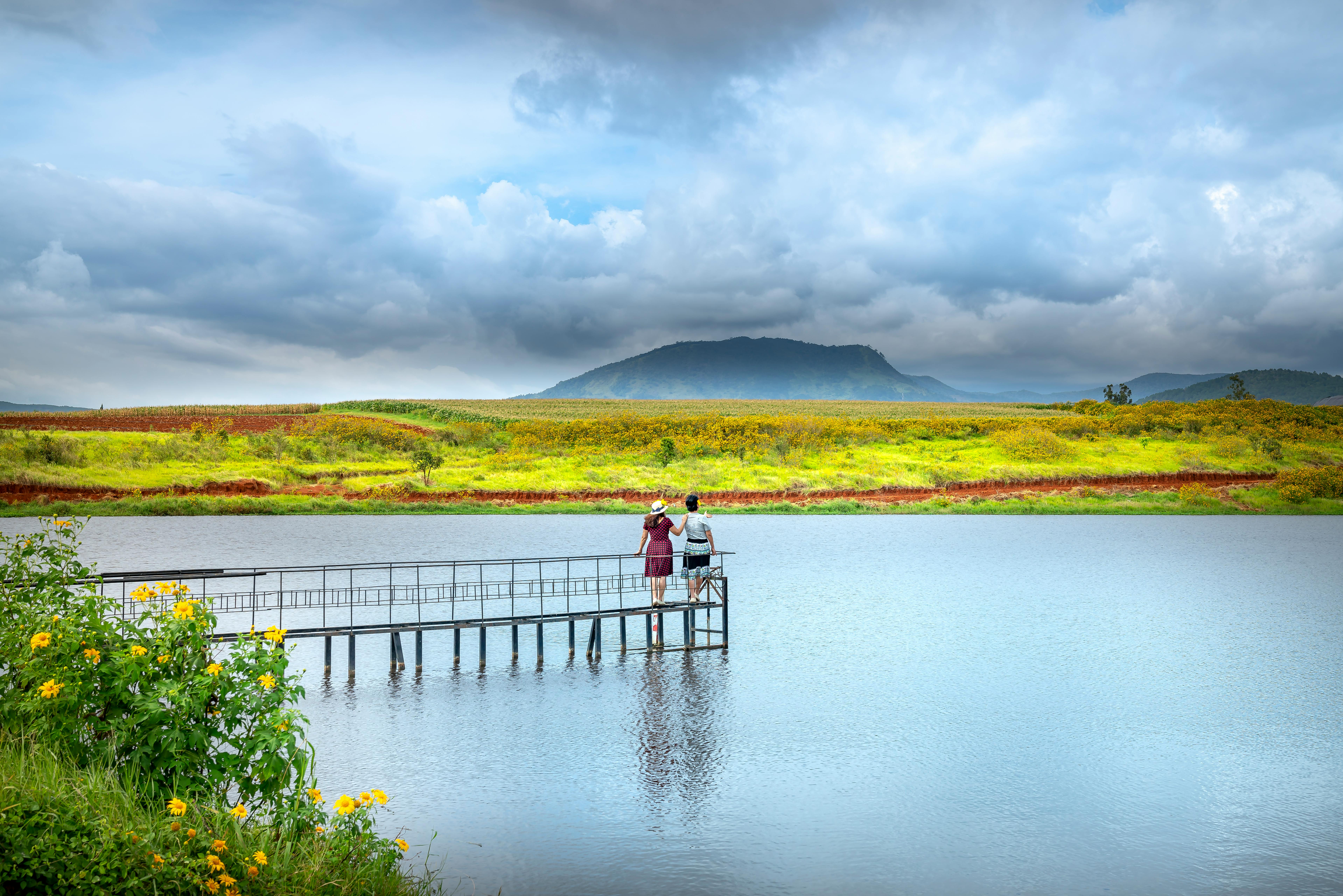 一对夫妇站在宁静湖泊旁的码头上，可以看见山脉和花海的景色