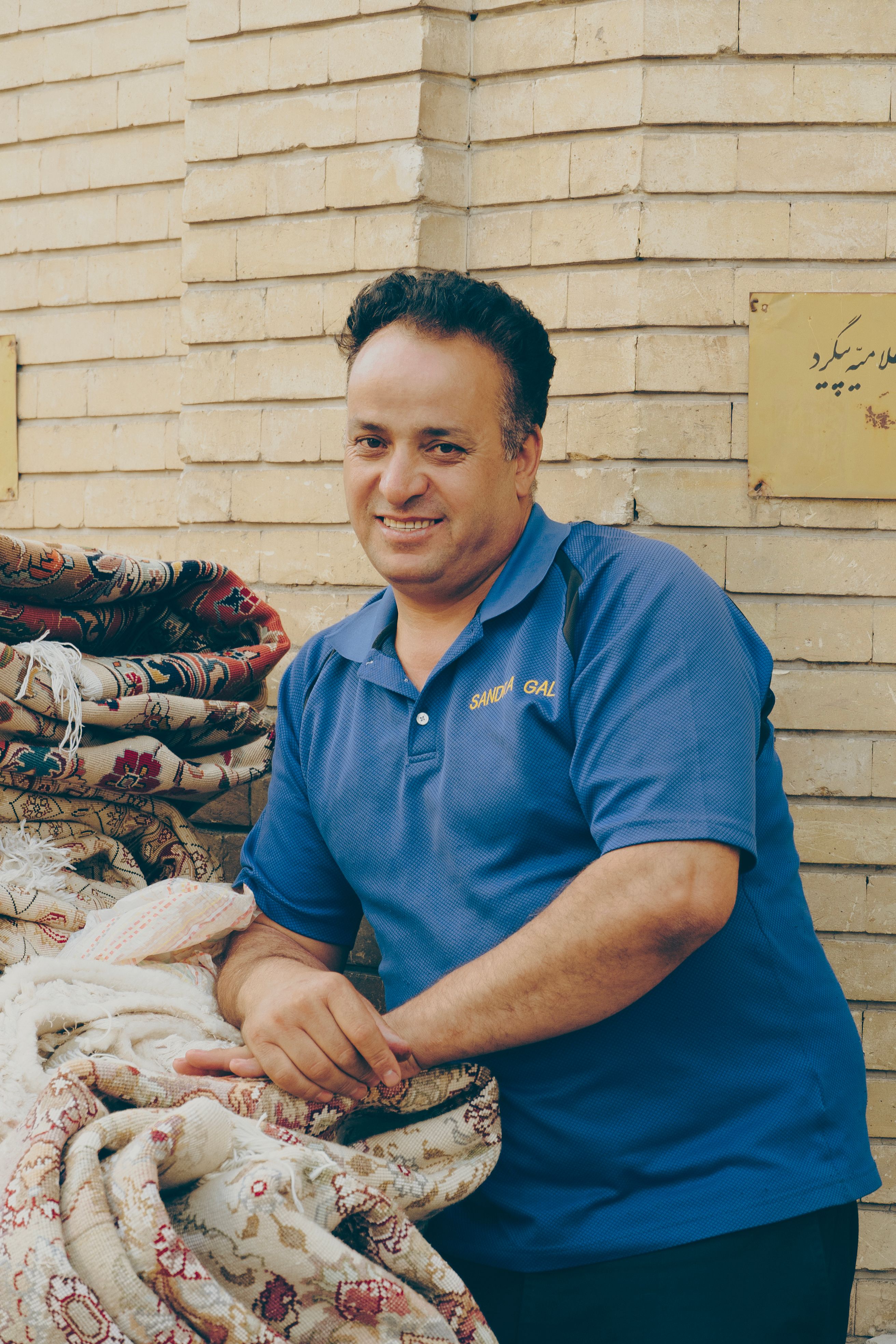 A smiling man in a blue polo poses with traditional rugs in Tabriz.