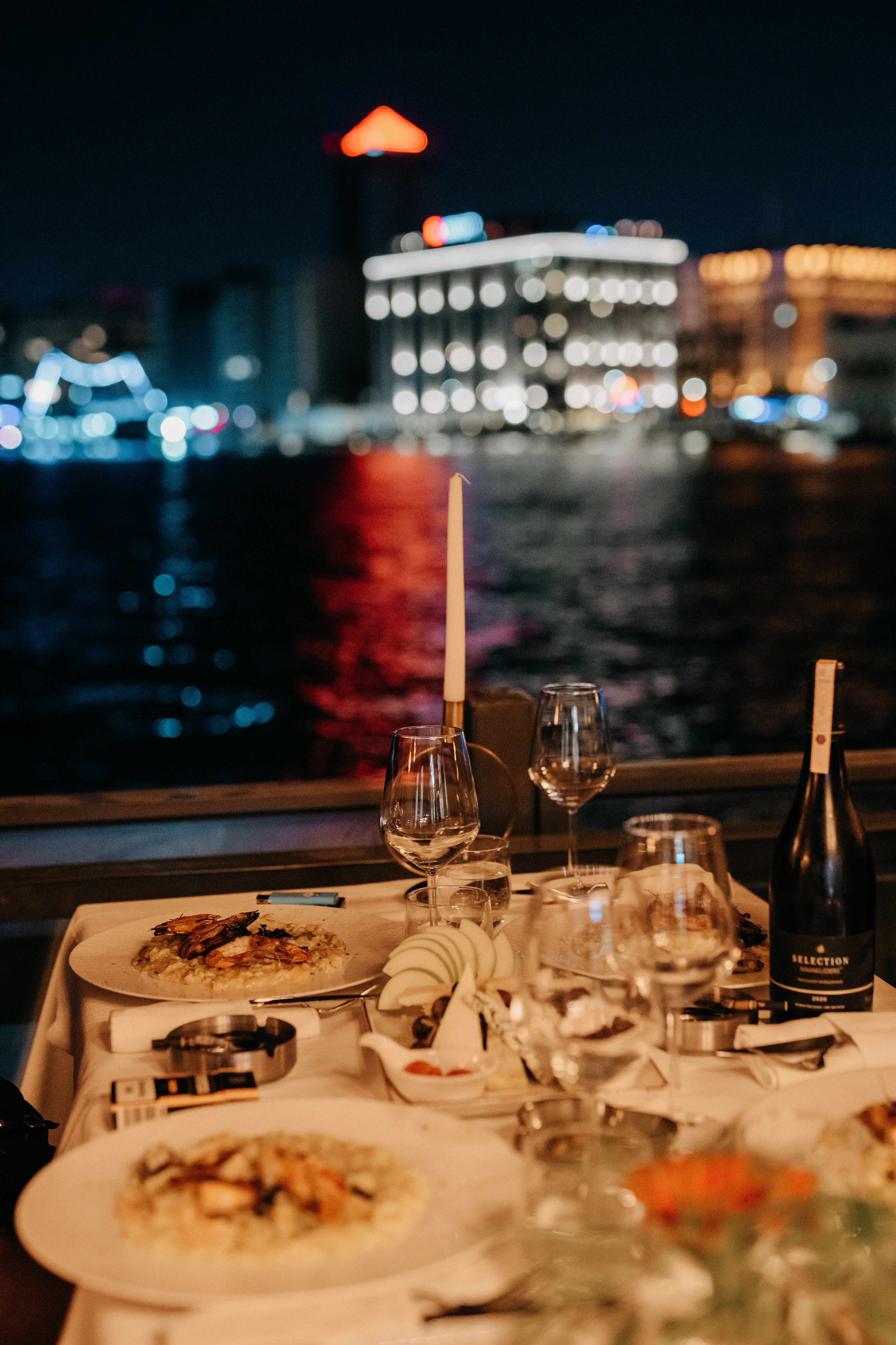 Enjoy a romantic dinner by the river at night, featuring elegant plates and a stunning cityscape view.