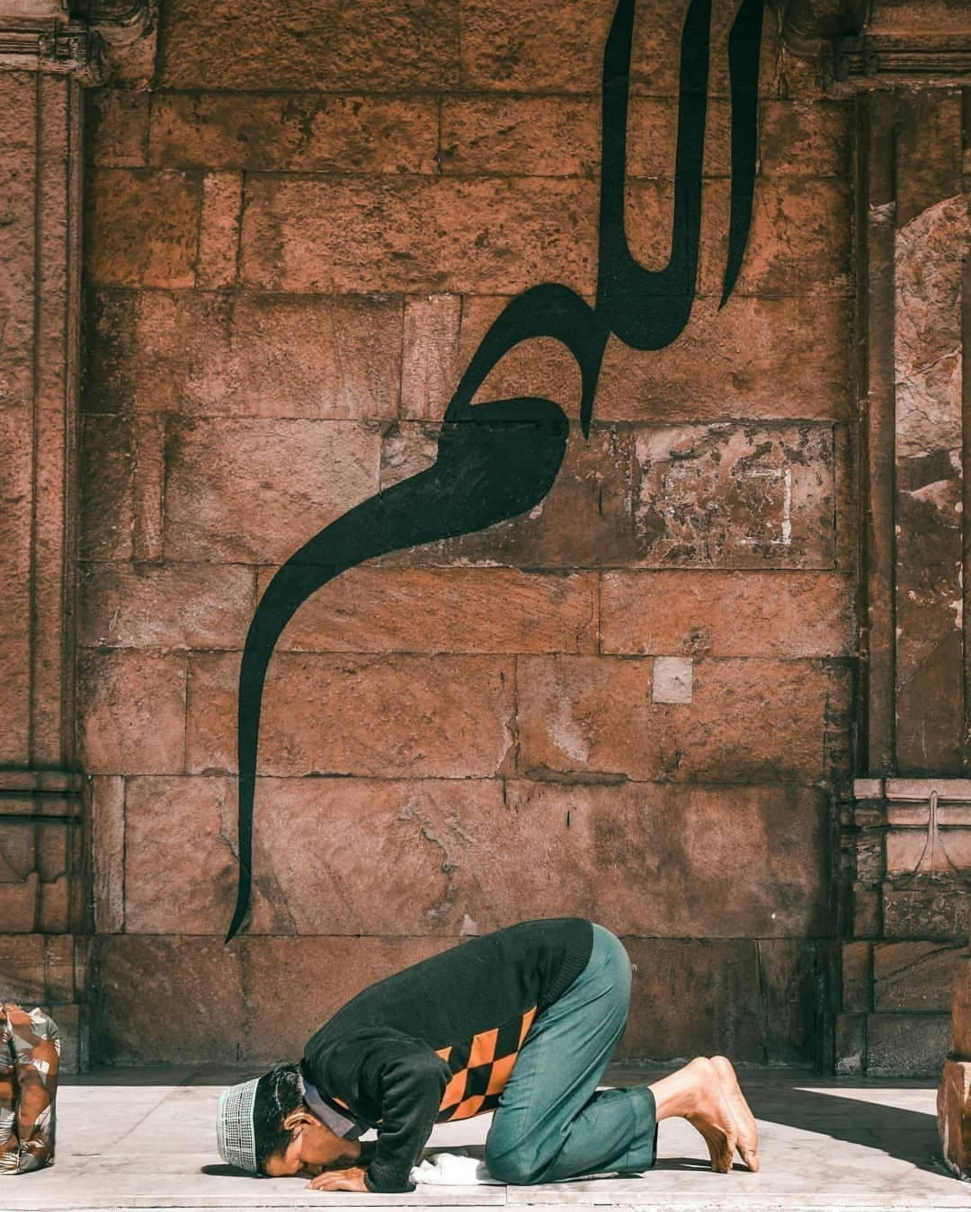 A man praying in a mosque, adorned with Arabic calligraphy on the wall.