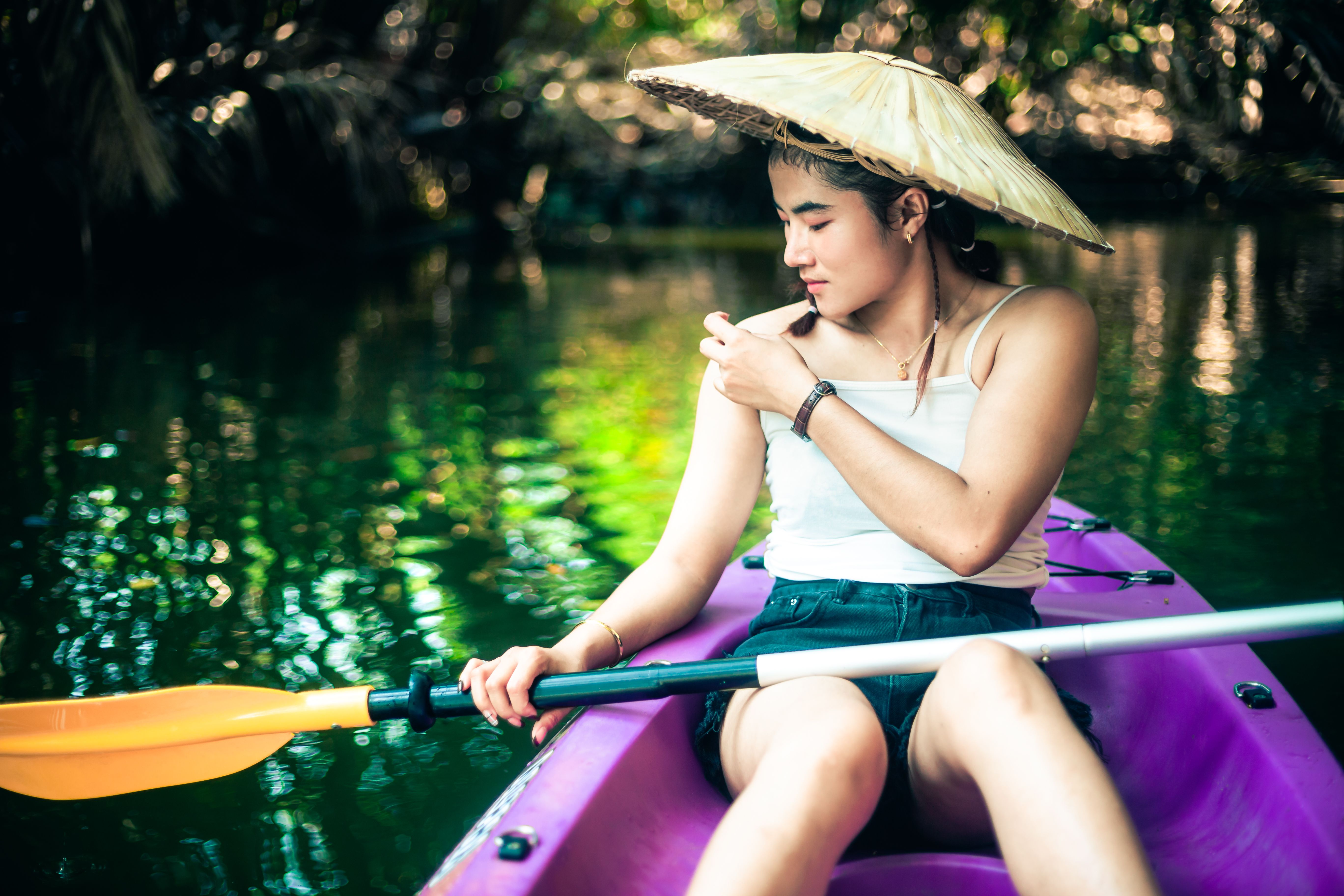 An Asian woman in a canoe with a conical hat paddles in peaceful waters surrounded by greenery.