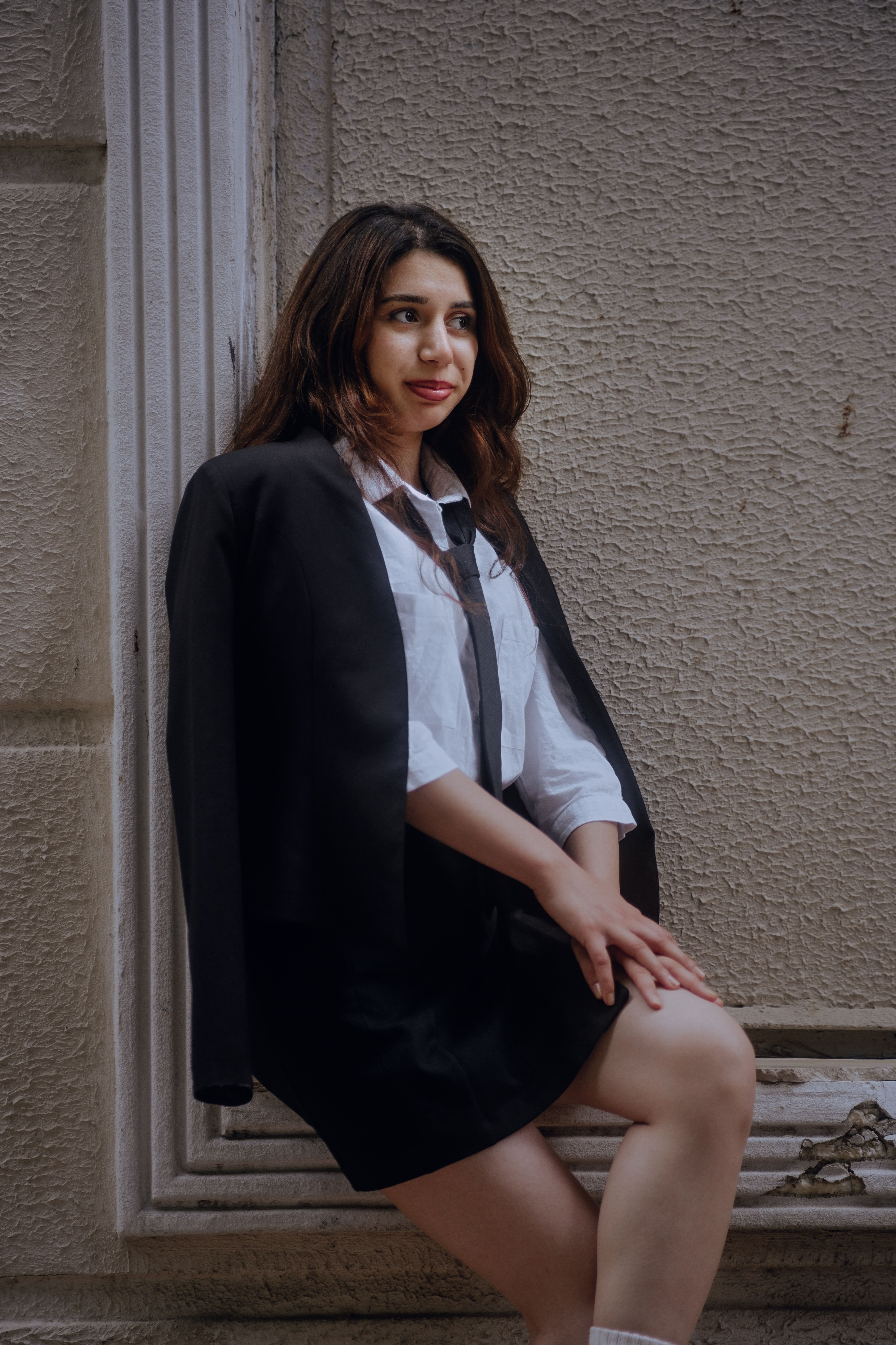 Portrait of a woman in a suit jacket and tie, sitting by a wall in an urban environment.