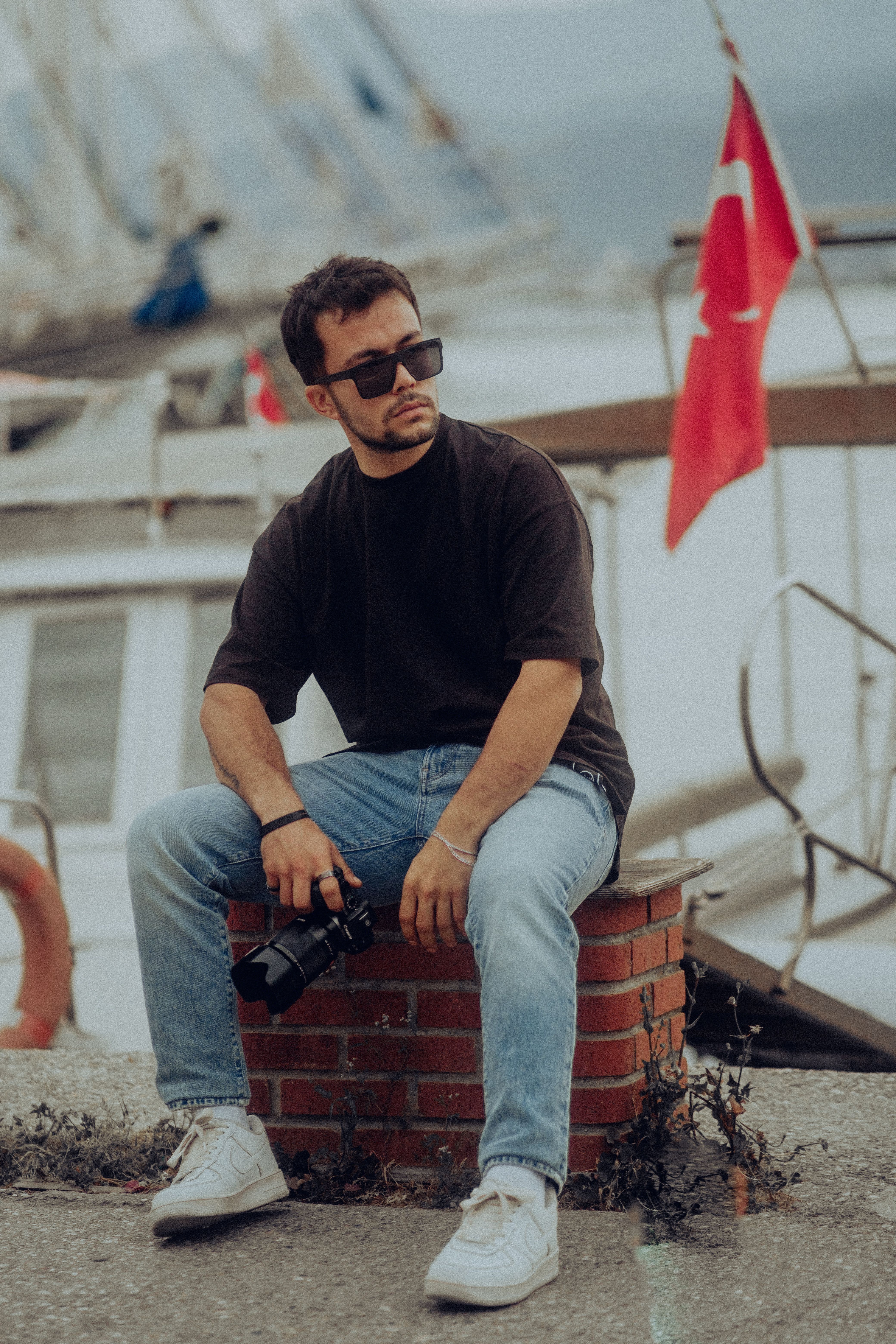 Stylish man in sunglasses sitting by a dock holding a camera, with Turkish flags in the background.