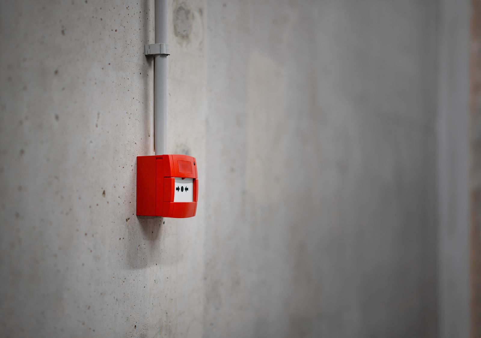 A small red fire alarm call point mounted on a rough grey concrete wall, its conduit running up out of frame, the rest of the wall empty