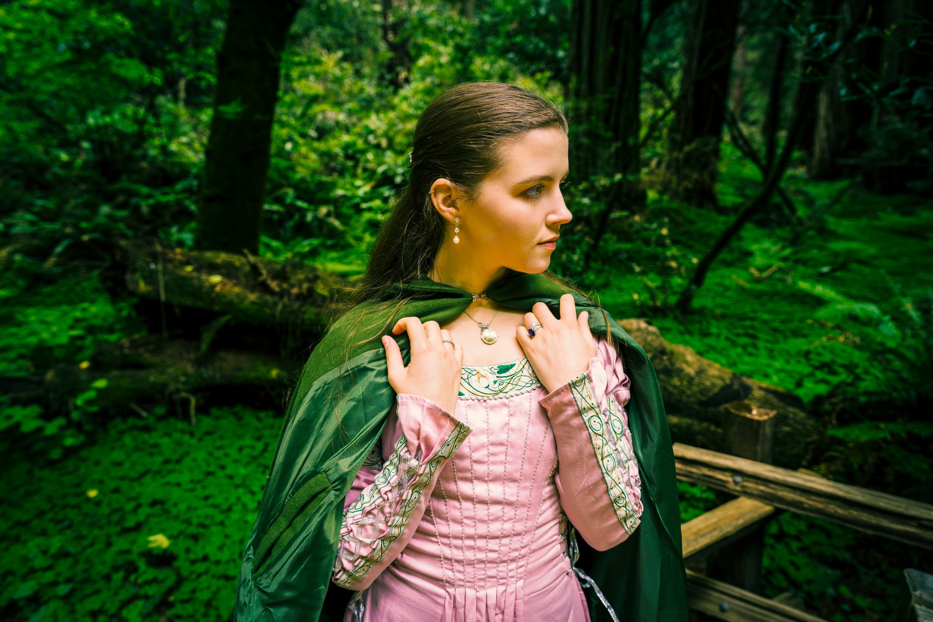 A woman in a pink dress and cloak stands in a lush forest, embodying a princess figure.