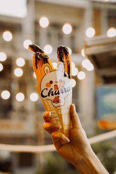 Close-up of a hand holding chocolate-dipped churros in a colorful cone outdoors.