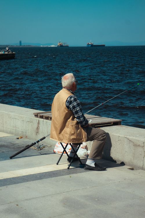Free Elderly man fishing by the sea on a sunny day at a coastal promenade. Stock Photo