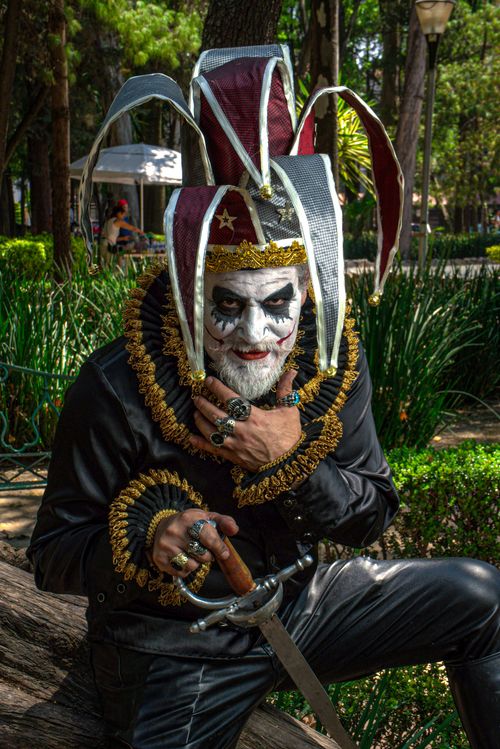 Free A man in a jester costume with makeup poses dramatically holding a sword in a park. Stock Photo
