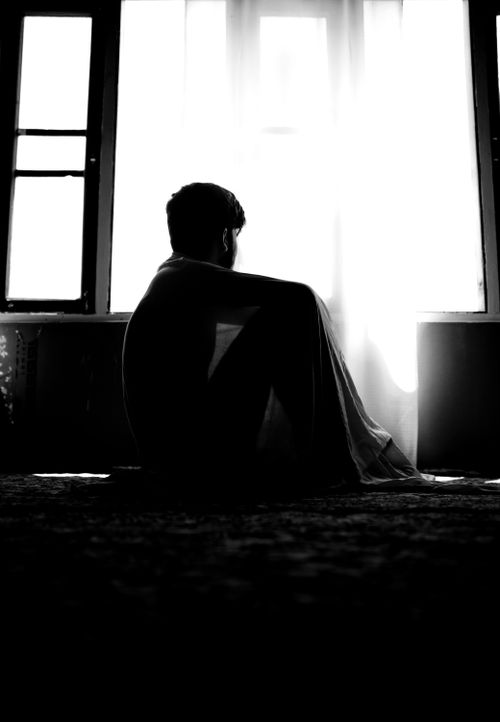 Free A silhouette of a person sitting and hugging their knees in front of a bright open window, creating a striking contrast. Stock Photo