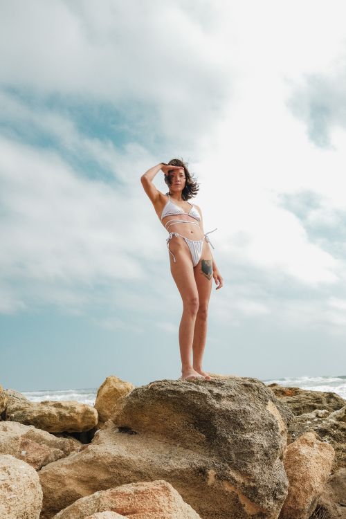 Free A woman poses confidently on a rock by the ocean in a bikini under a bright sky. Stock Photo