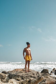 A muscular man stands confidently on a rocky beach, gazing at the ocean.
