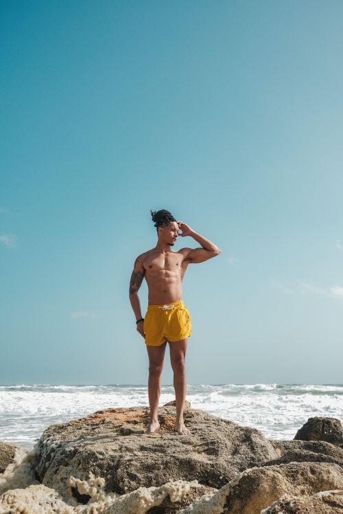 Free A fit man in yellow shorts poses on rocky beach under clear sky. Stock Photo