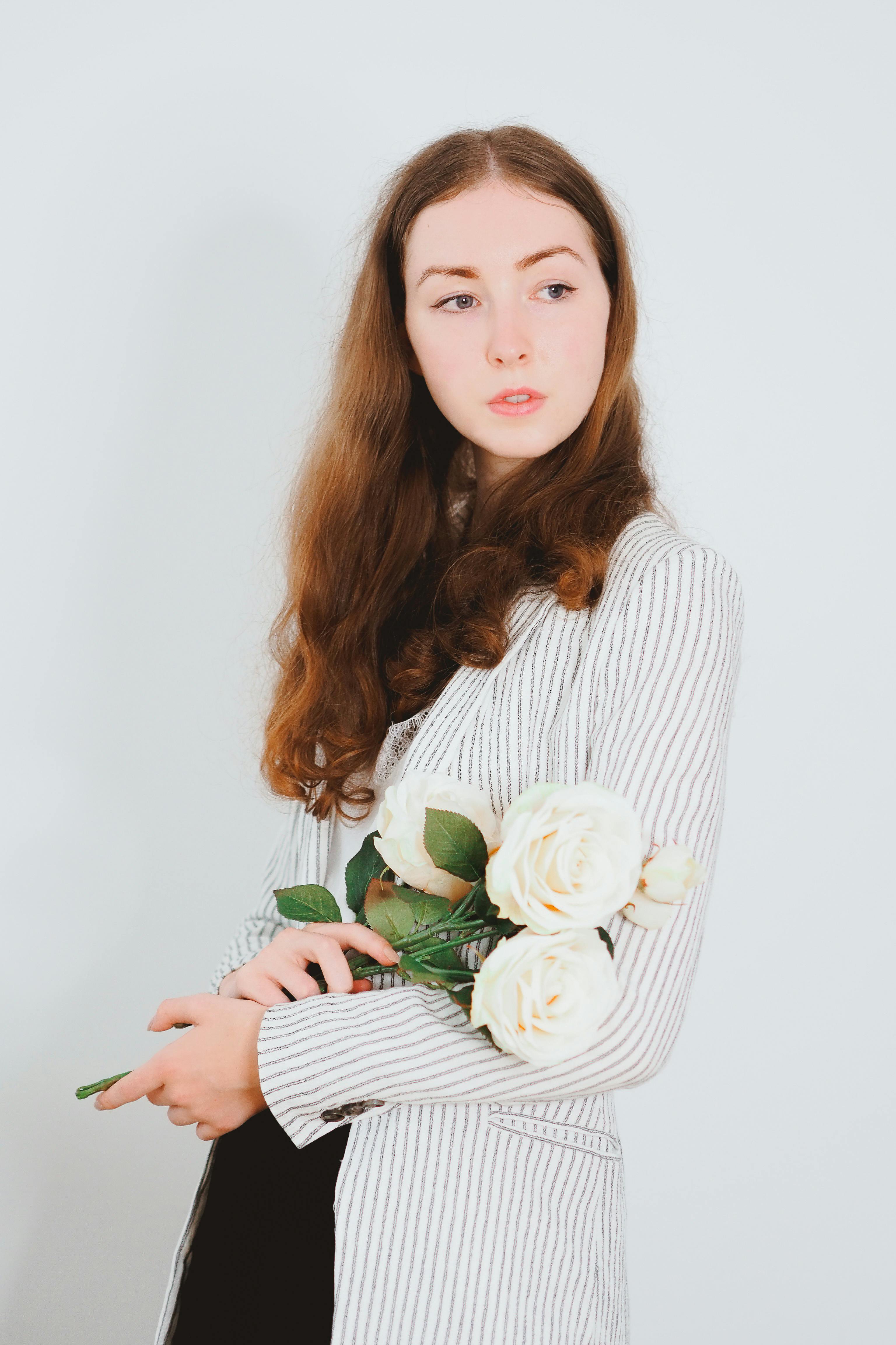 Fashion portrait of a redhead woman in a striped blazer holding white roses in a studio setting.