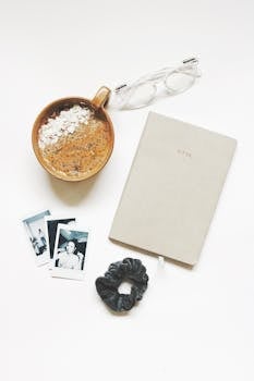 A cozy flatlay featuring a coffee, notebook, eyeglasses, and vintage photos on a white background.