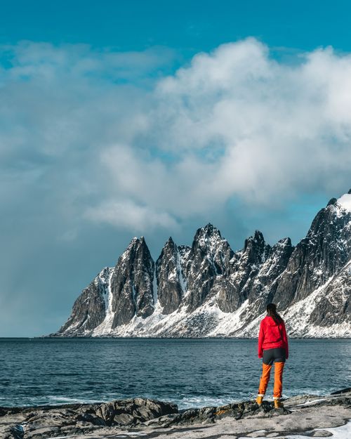 Free A lone woman gazes at snow-capped mountains along a chilly seashore, capturing winter's serene beauty. Stock Photo
