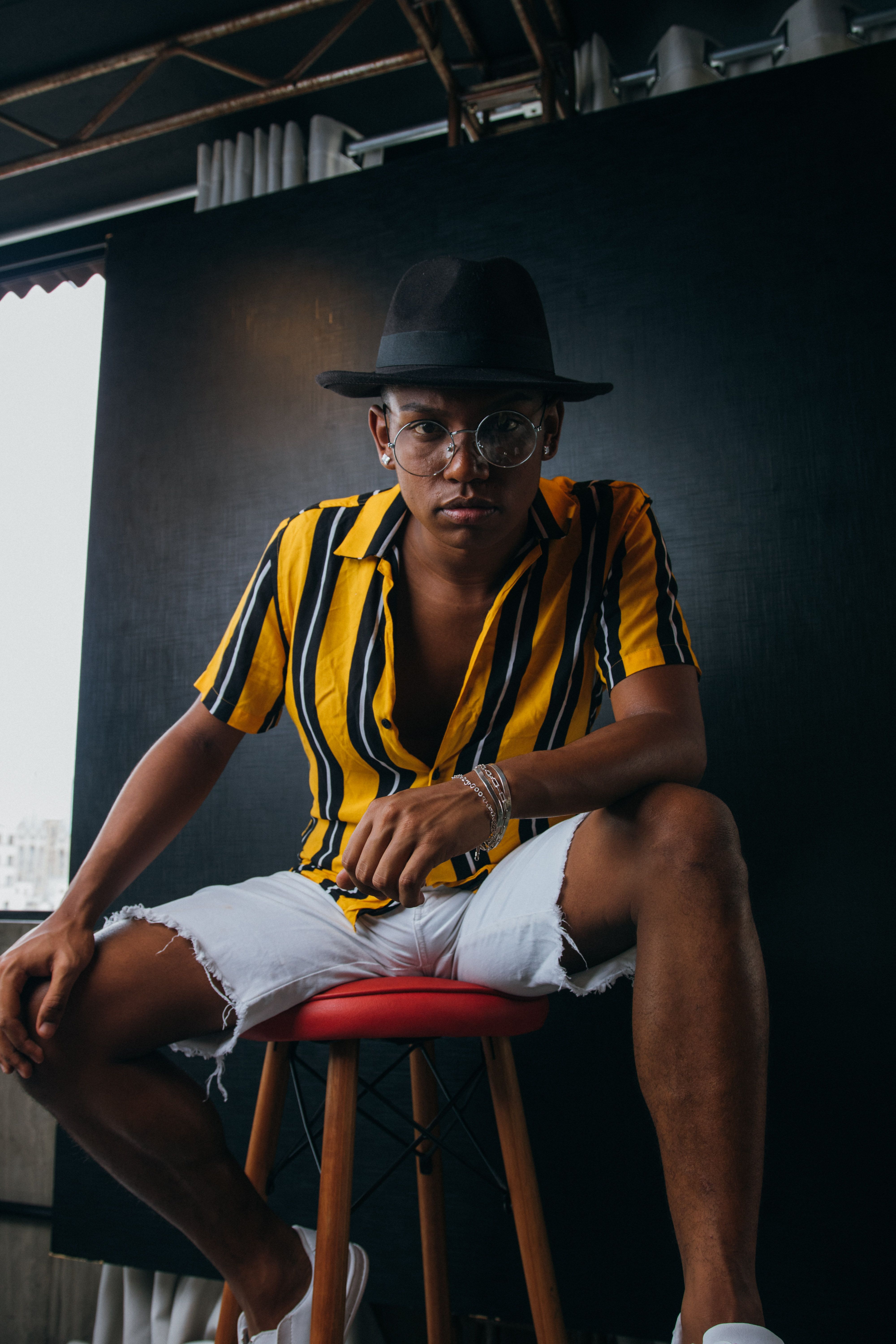 Fashionable adult wearing yellow striped shirt and fedora hat, seated on a stool indoors.