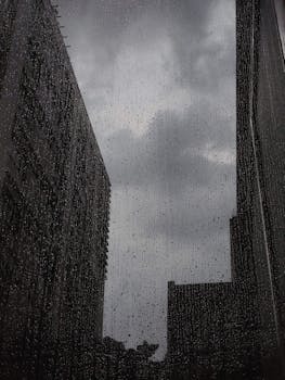 A dark urban scene with rain droplets on a window overlooking tall buildings.