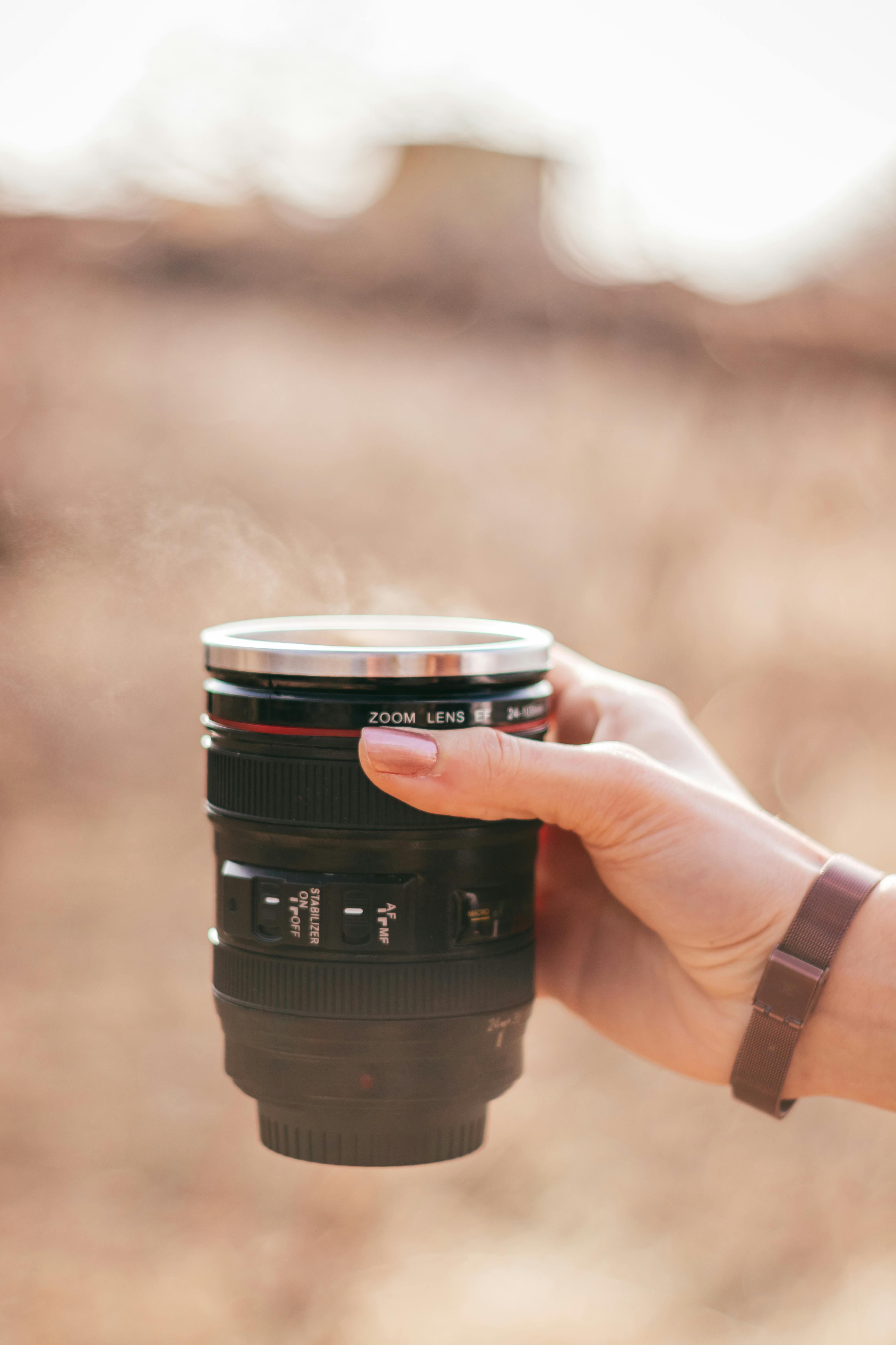A person holding a mug that resembles a camera lens, with a warm outdoor background.