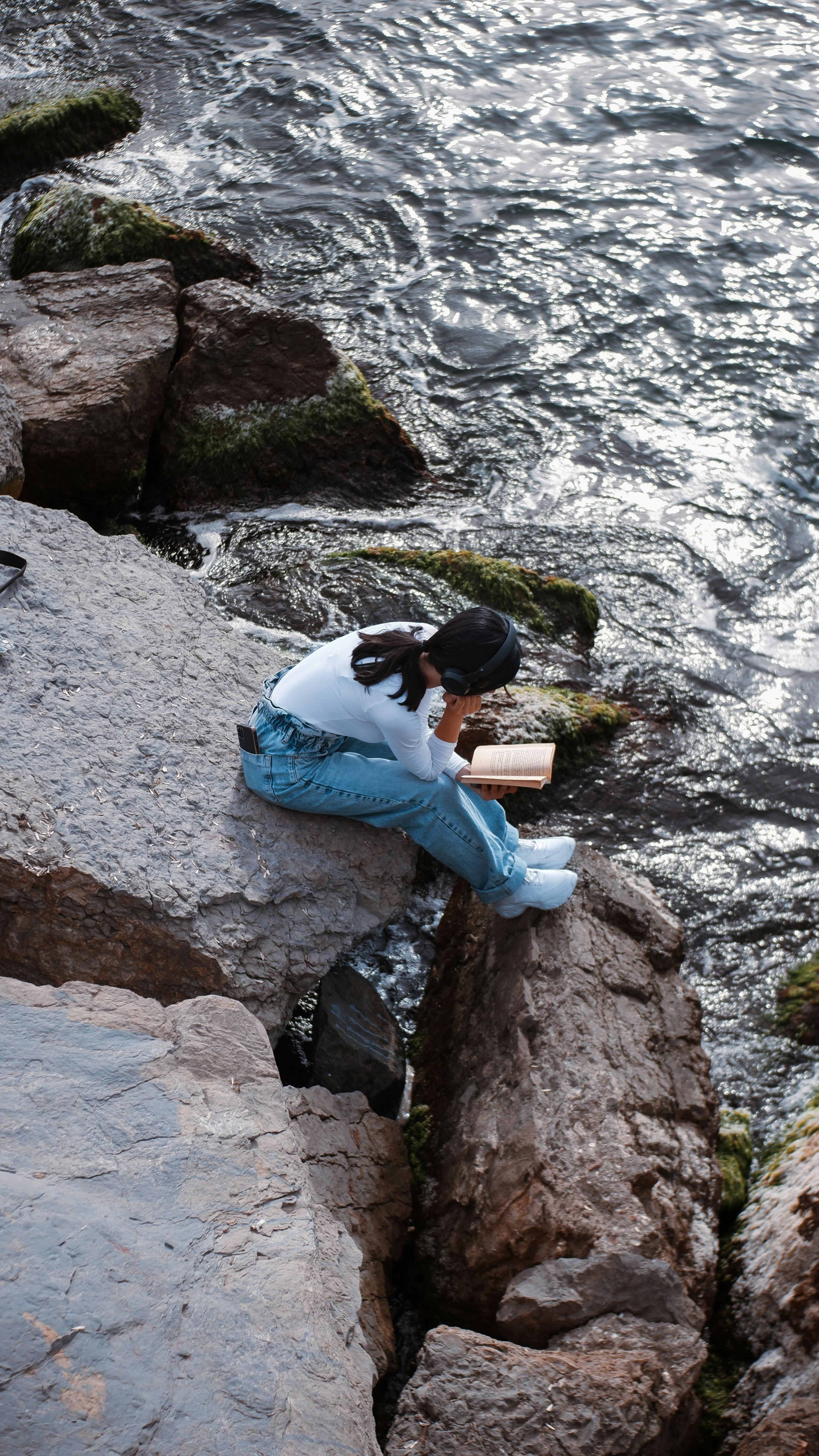 A woman enjoys reading a book while sitting on rocky coastal terrain