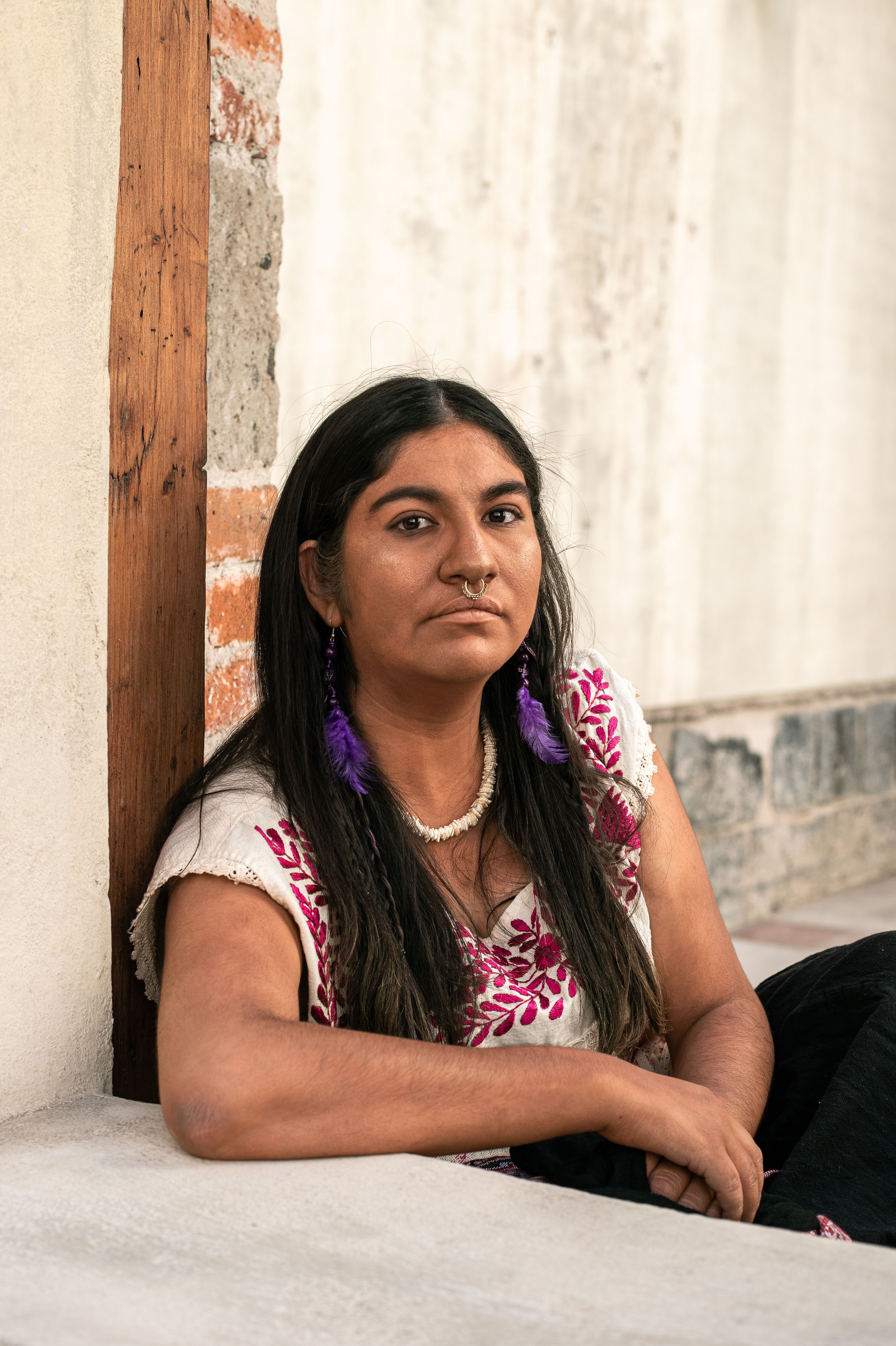 Portrait of a woman in traditional clothing, showcasing cultural jewelry and style, seated against a wall.