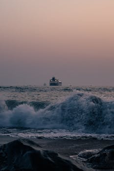 Dramatic seascape with cargo ship at sunset, featuring crashing waves and misty horizon.
