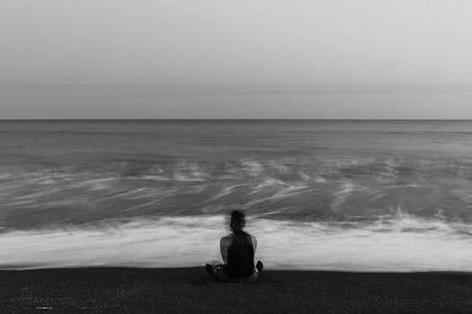 A contemplative scene of a lone figure sitting on a beach, facing the ocean in a black and white composition.