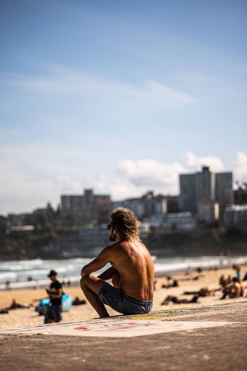 Free Tanned man sitting shirtless at Sydney Bondi Beach, overlooking the sea and city skyline. Stock Photo