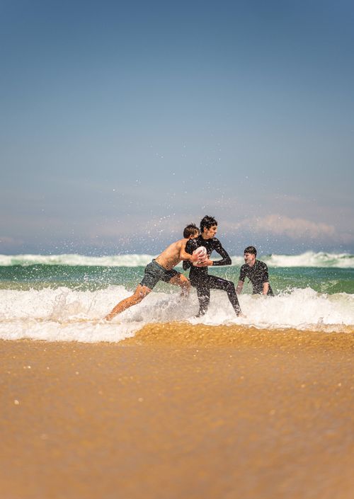 Free Three young men enjoying a game of rugby on a sunny Sydney beach with crashing waves. Stock Photo