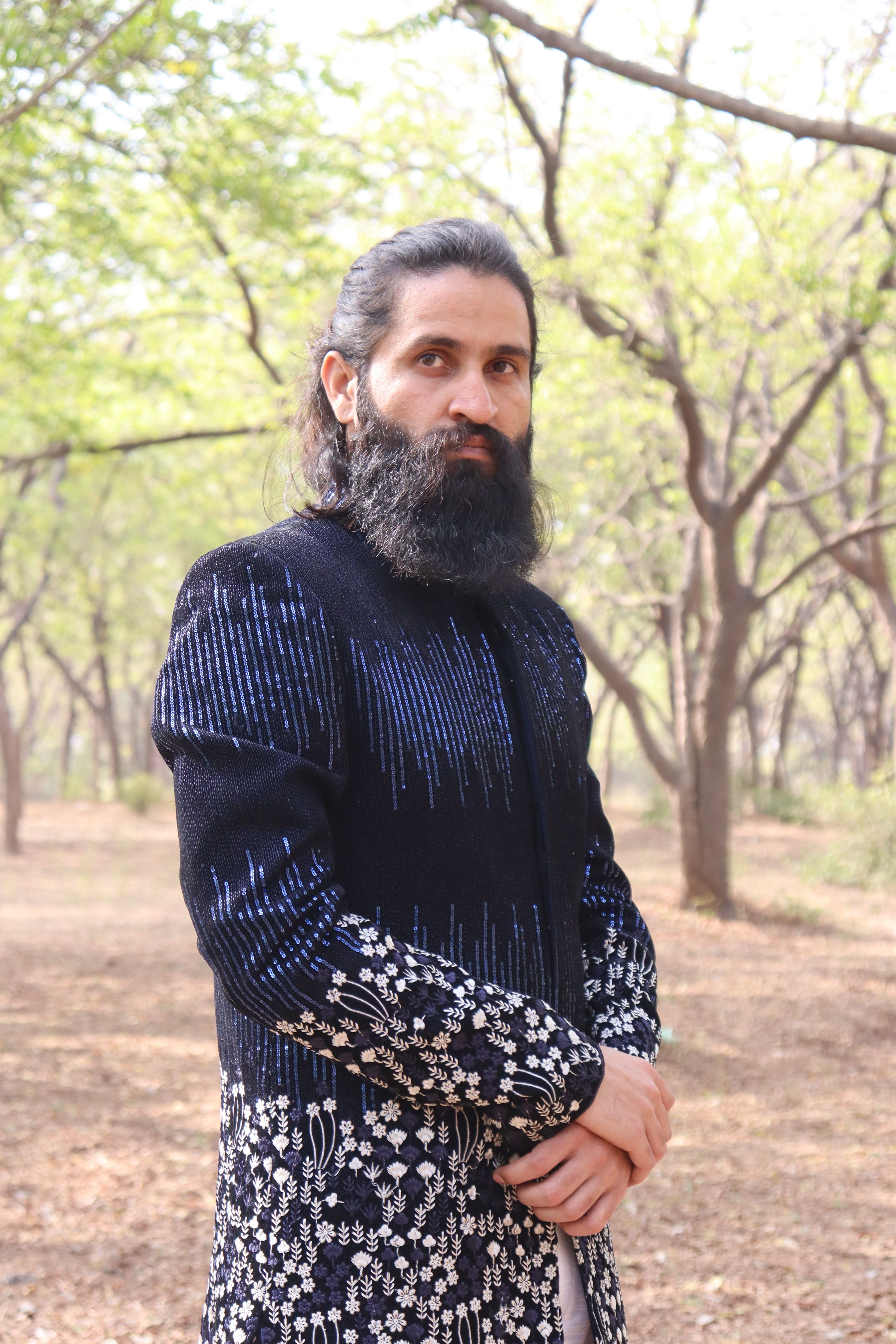 A bearded man in traditional clothing poses in a serene forest setting during daylight.