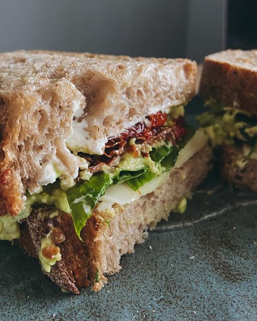 Free Close-up of a delicious sandwich with avocado, sun-dried tomatoes, and leafy greens on rustic bread. Stock Photo
