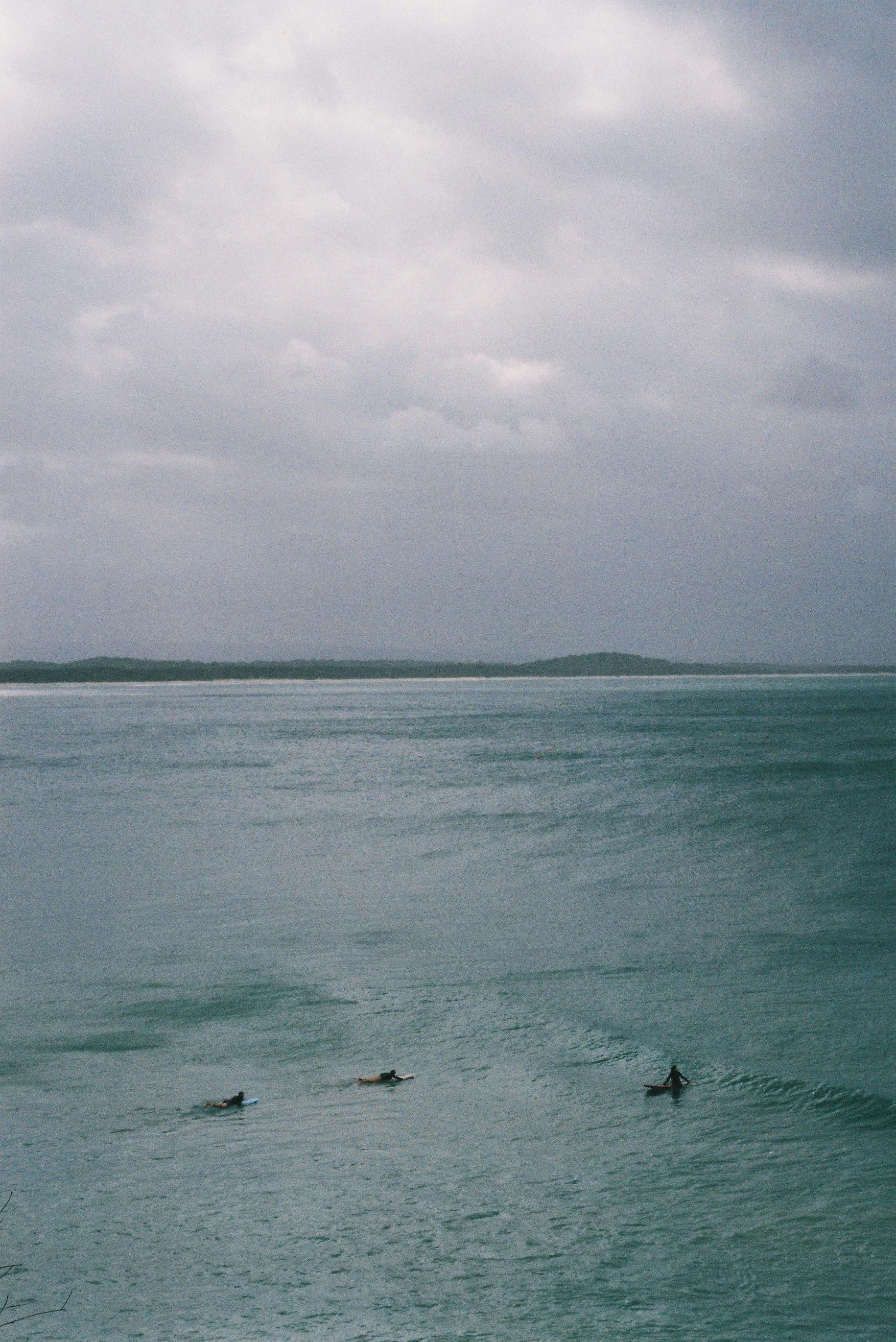 A serene aerial view of three surfers paddling on a calm ocean under a cloudy sky.