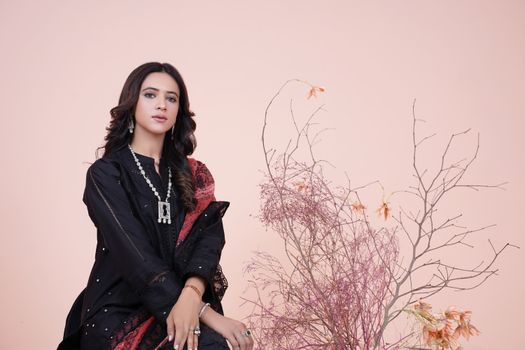 A stylish woman in a black dress poses with floral branches, set against a minimalist pink studio background.