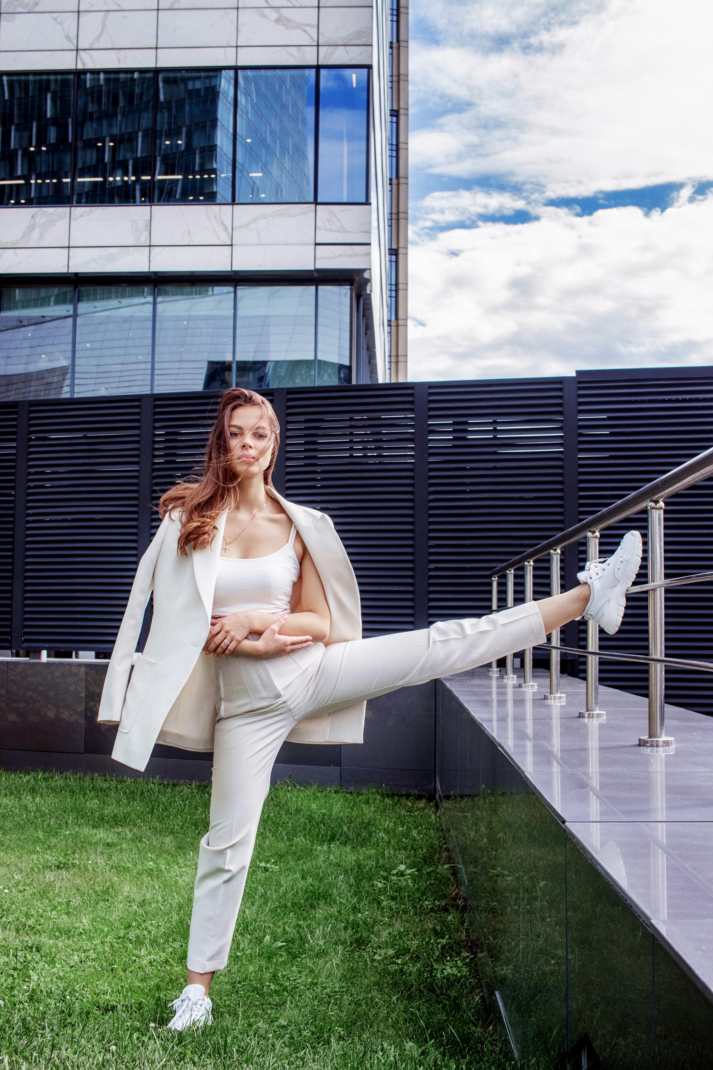 A young woman in stylish attire stretching her leg outdoors in a modern urban area.